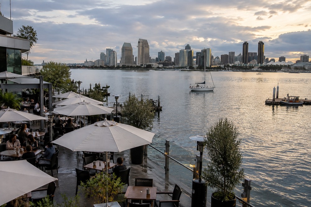 Outdoor dining terrace beside calm bay water with city towers in the distance