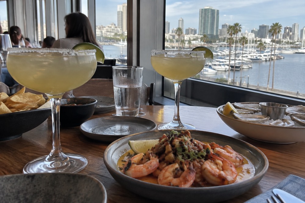 Table with margarita glasses and seafood dishes beside a window overlooking a marina