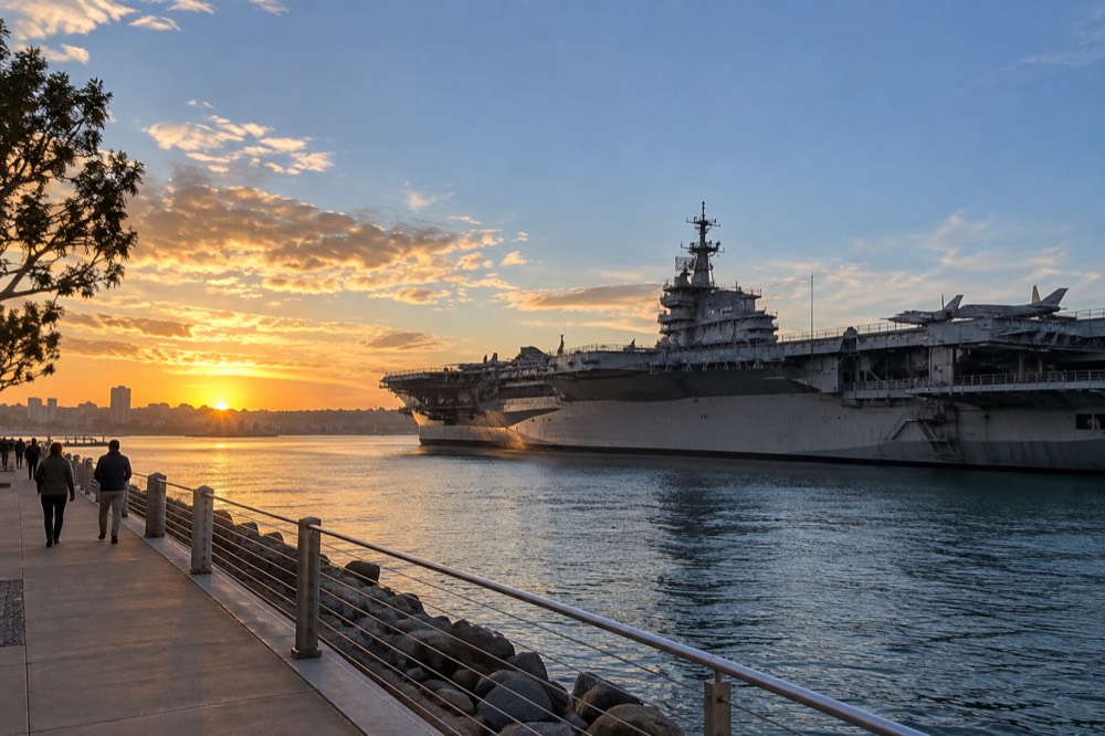 A waterfront promenade at golden hour with a docked aircraft carrier on the far side of the bay