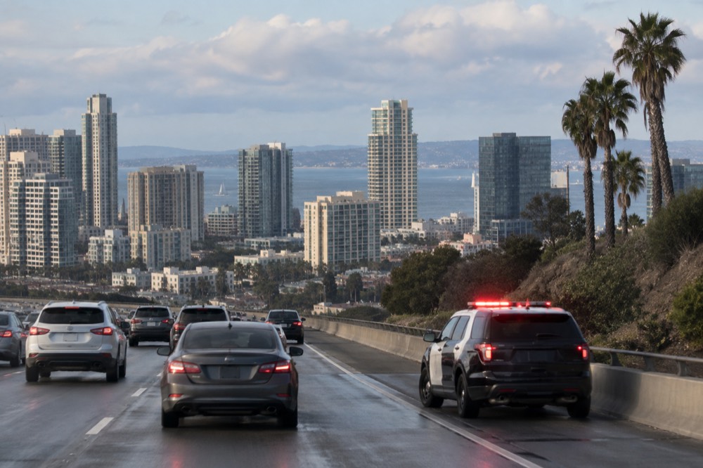 A highway patrol vehicle parked on a freeway shoulder with red lights flashing beside slowed traffic