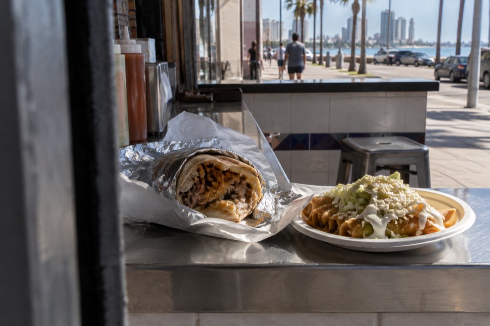 A California burrito wrapped in foil beside a plate of rolled tacos on a taqueria counter