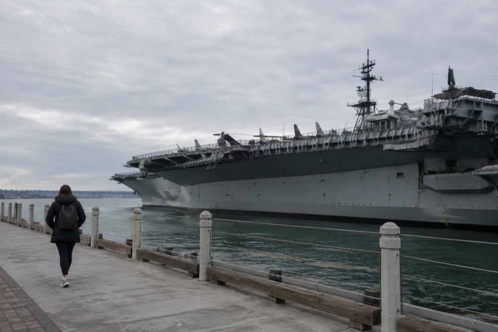 A visitor walking along the Embarcadero with the USS Midway ship in the background