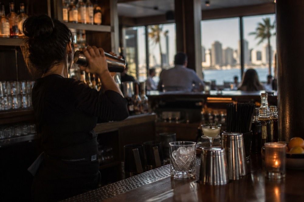 A bartender shaking a cocktail at a low-lit wooden bar with glassware on the counter