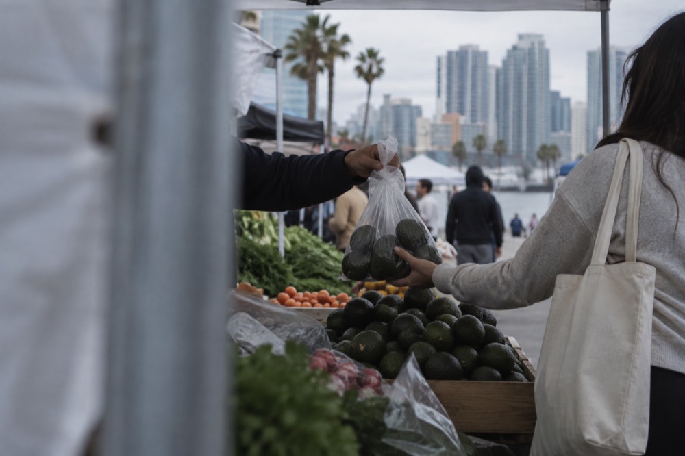 A vendor handing a bag of avocados to a shopper at a market stall
