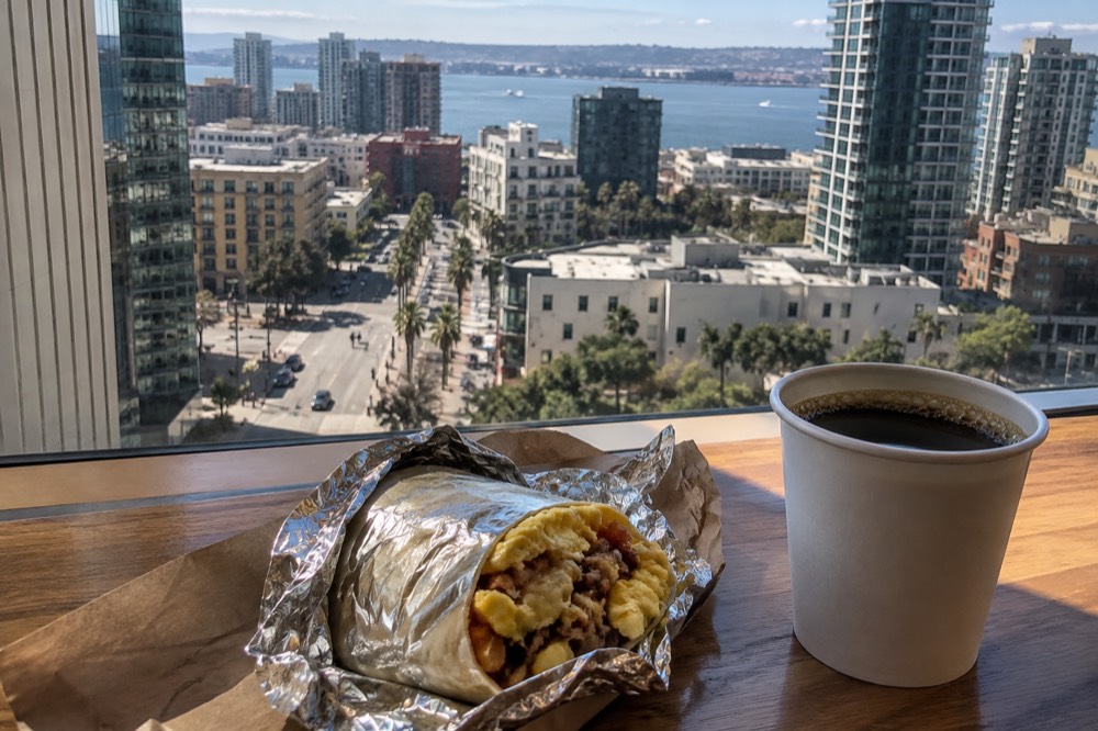 A breakfast burrito wrapped in foil on a wooden counter beside a coffee cup