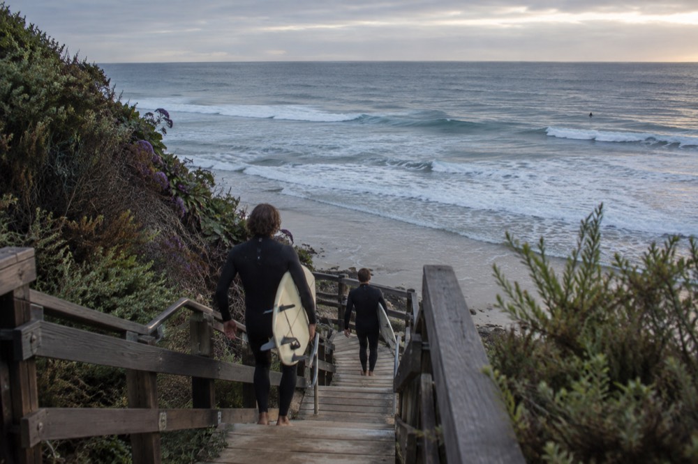 Surfers walking down a wooden beach staircase toward the ocean in the early morning