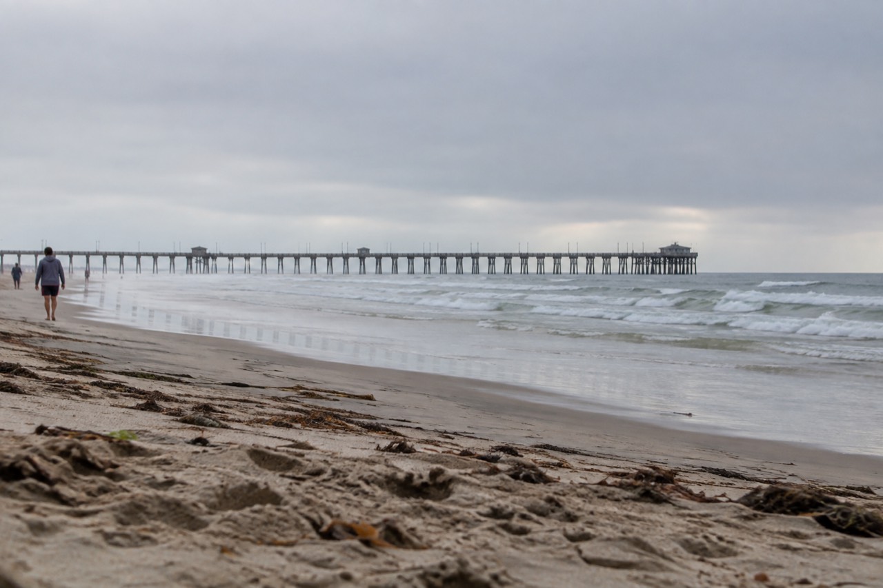A wide sandy beach with a long pier reaching out over low waves under a pale morning sky
