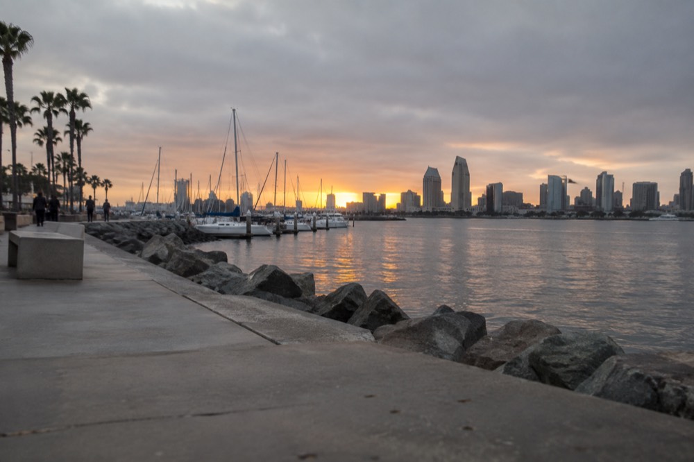 Waterfront promenade at sunset with masts of sailboats and a city skyline across the bay