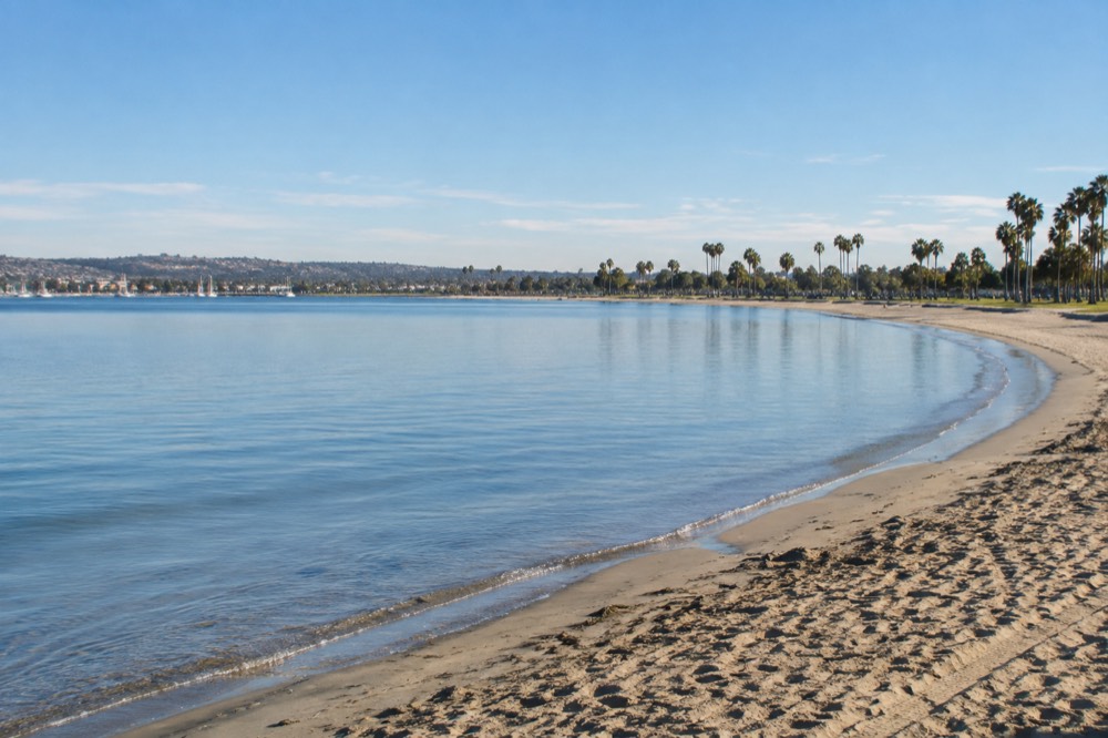 Calm bay water at sunrise with a long curve of sandy shoreline and distant palm trees