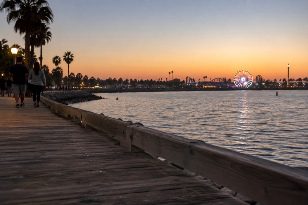 A boardwalk beside the bay at dusk with amusement park lights glowing in the distance