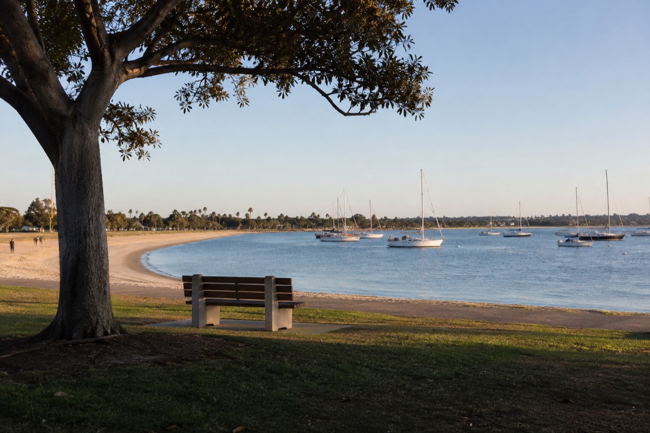 Calm blue bay water with sailboats and a curved shoreline park under a clear sky