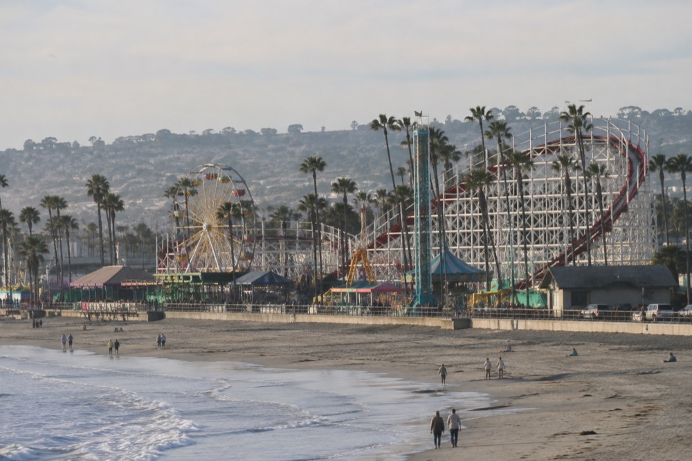 A wooden roller coaster and amusement park rides standing beside a beach boardwalk