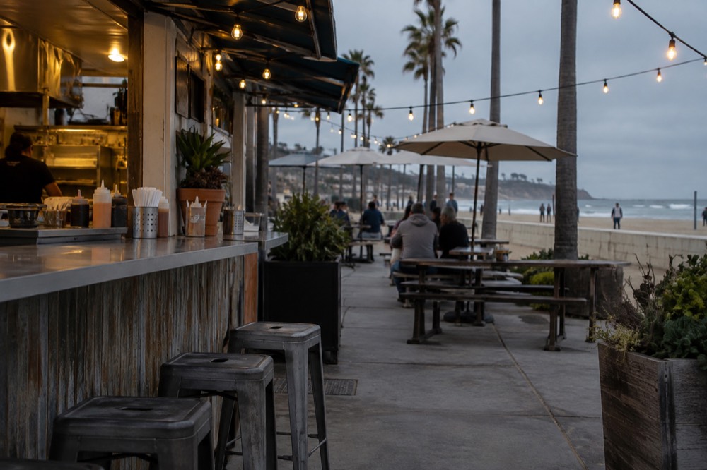 An outdoor taco shop counter with string lights and patio tables in the evening
