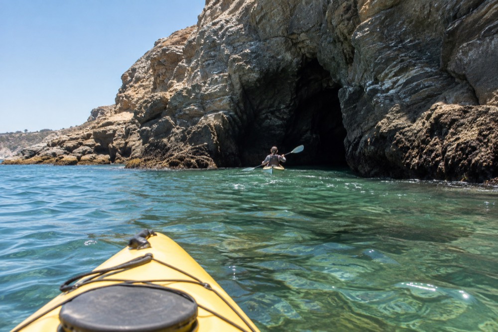 A kayak paddling across clear green water toward a rocky sea cave opening