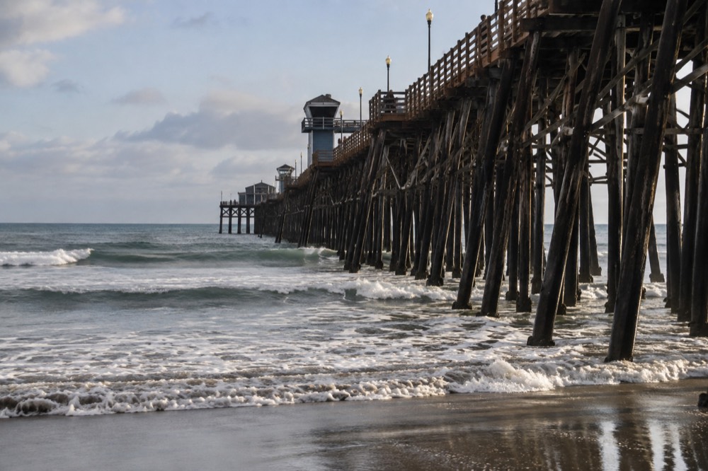 A long wooden pier extending over a wide sandy beach with small waves rolling toward the shore