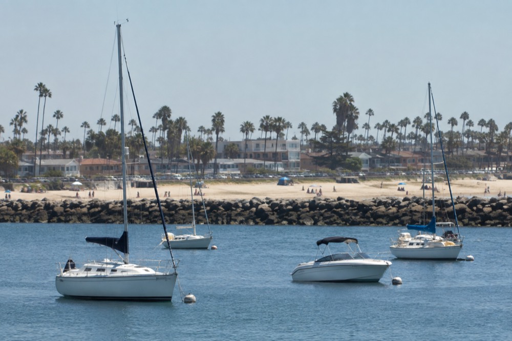 Small craft moored in a protected harbor with a sandy beach and low buildings along the shore