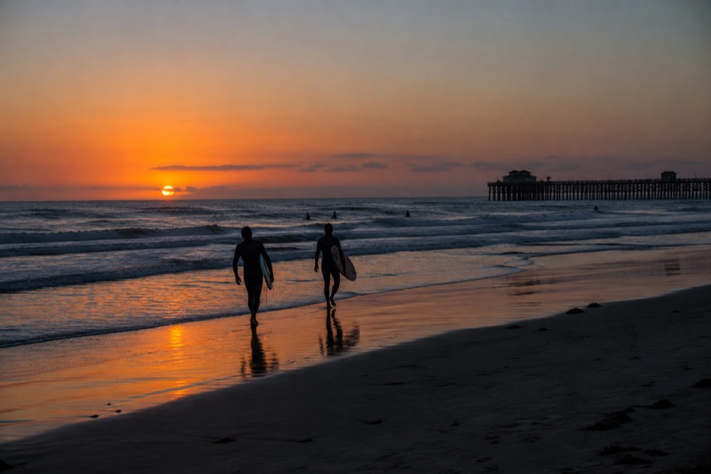 Golden sunset light over open ocean with silhouetted surfers walking along wet sand