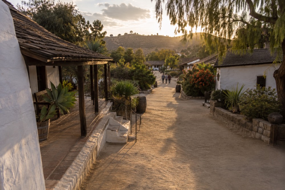 Historic wooden buildings along a walkway with distant hills at golden hour