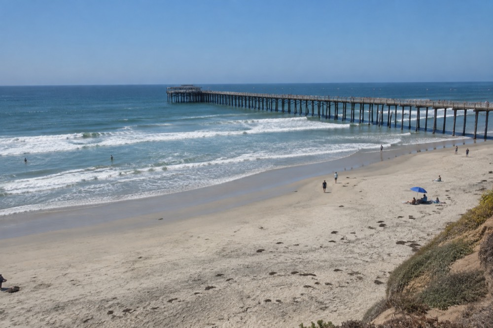 Wide sandy beach with a long wooden pier extending over gentle waves under a clear sky