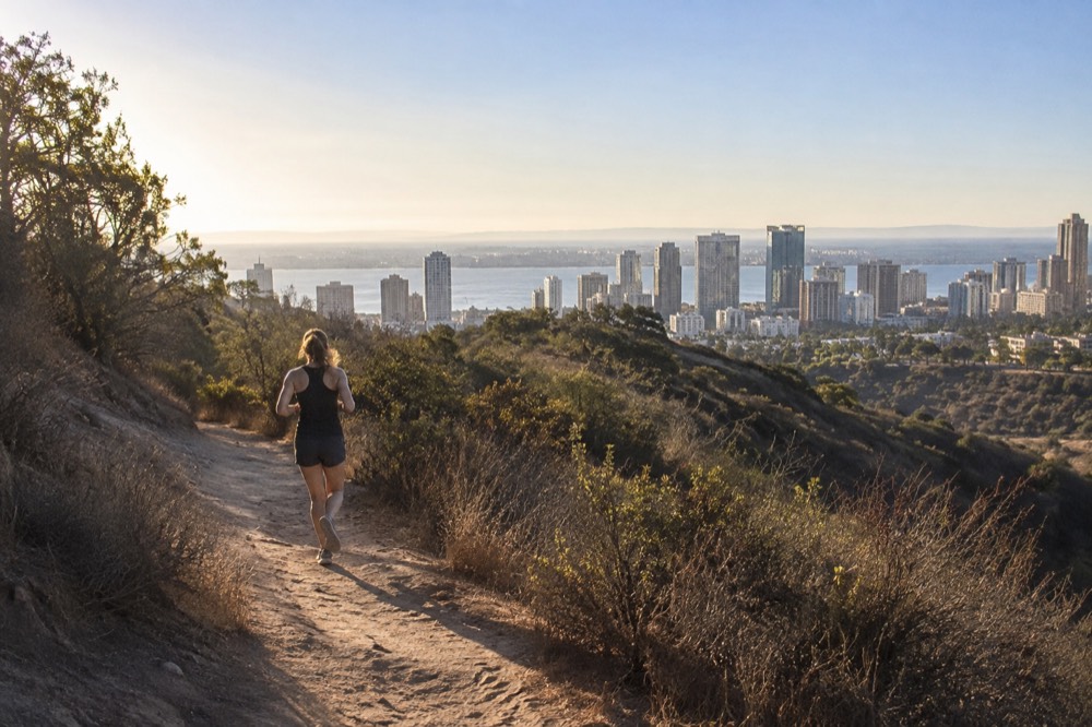 Dirt trail through scrub covered canyon with city buildings visible beyond