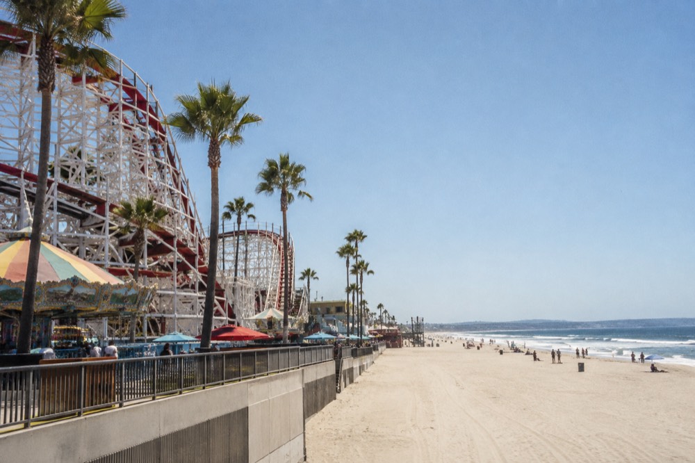 A wooden roller coaster and boardwalk rides beside a wide sandy beach under a blue sky