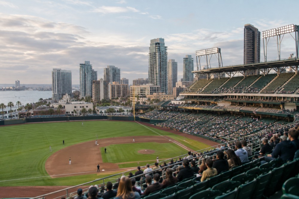 A baseball stadium interior with green seats, a scoreboard and city buildings visible beyond the outfield