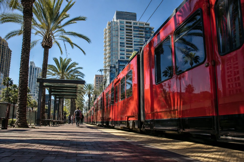A red trolley car at a downtown station with palm trees and buildings behind it