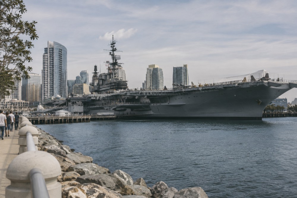 A large gray aircraft carrier museum moored at a waterfront pier with a city skyline behind it