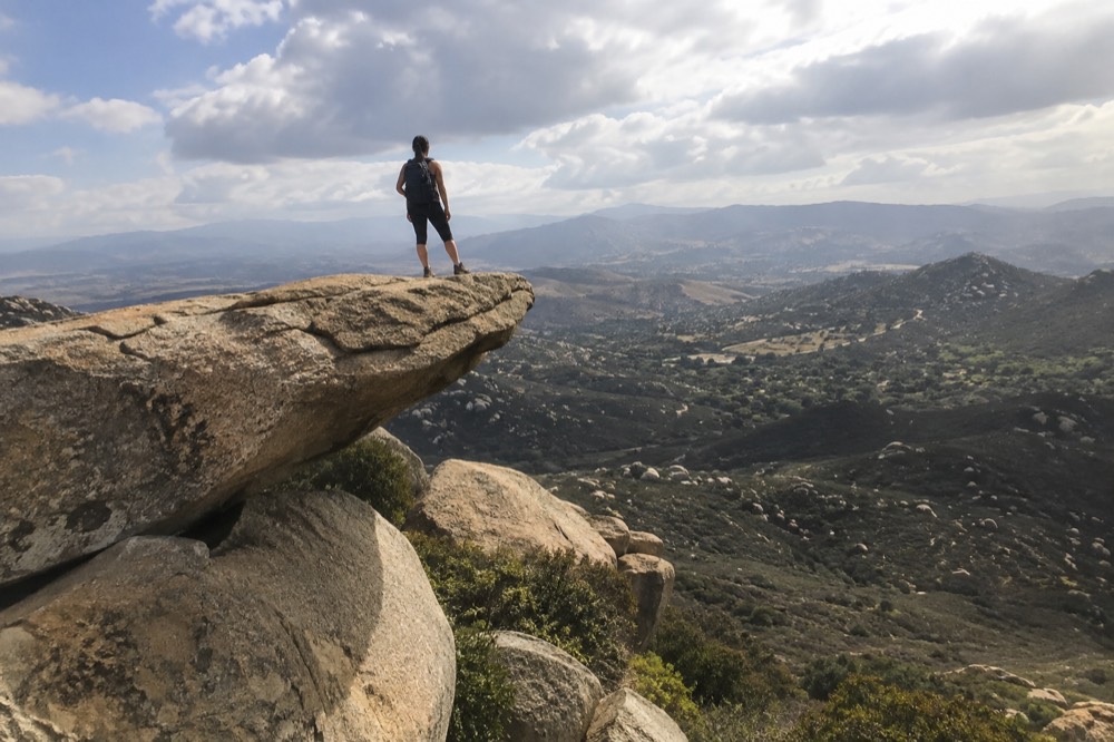 A hiker standing on a narrow rock ledge above a wide valley of low hills
