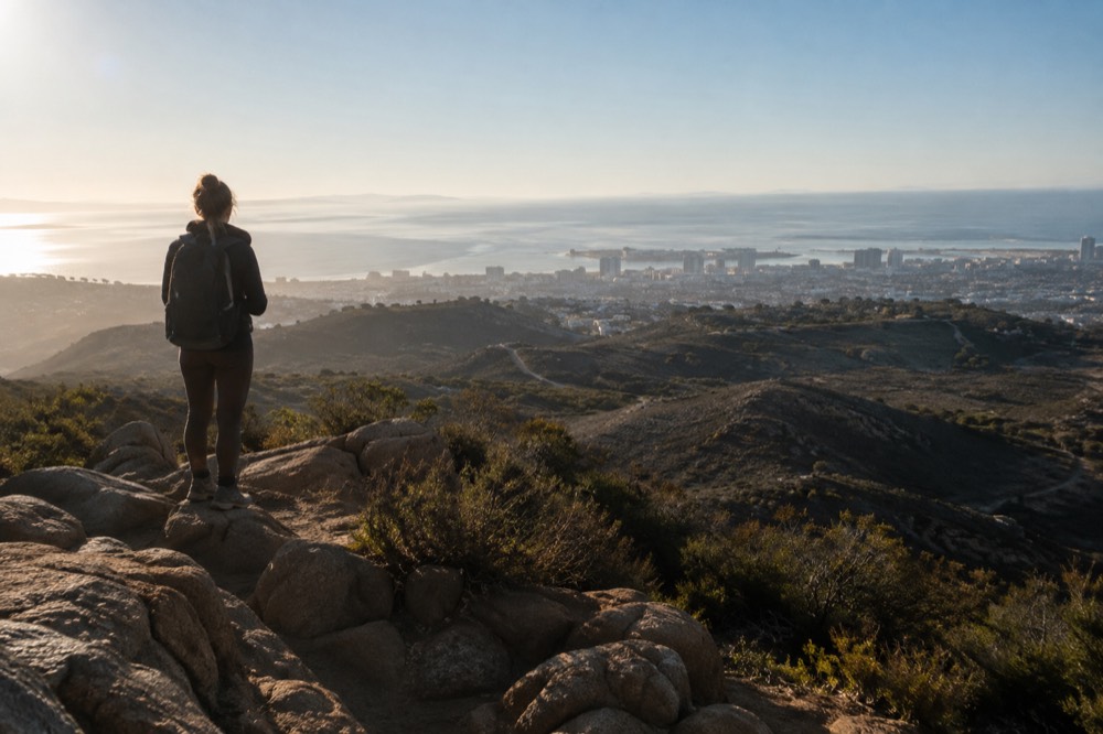 A hiker looking out from a rocky summit over rolling hills toward a distant ocean haze