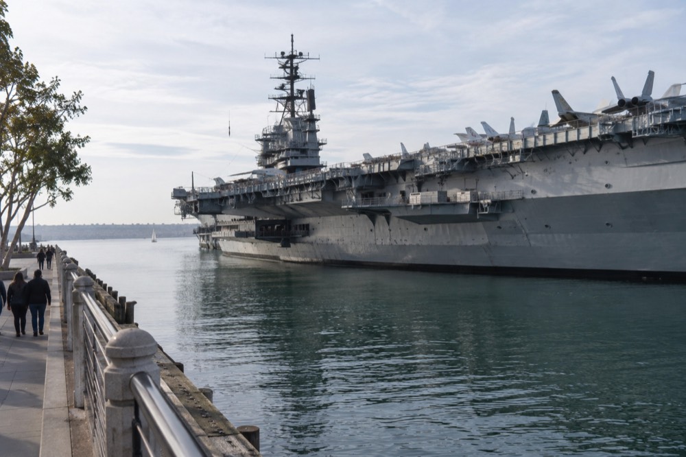 A calm harbor waterfront with a large gray aircraft carrier moored beside a promenade