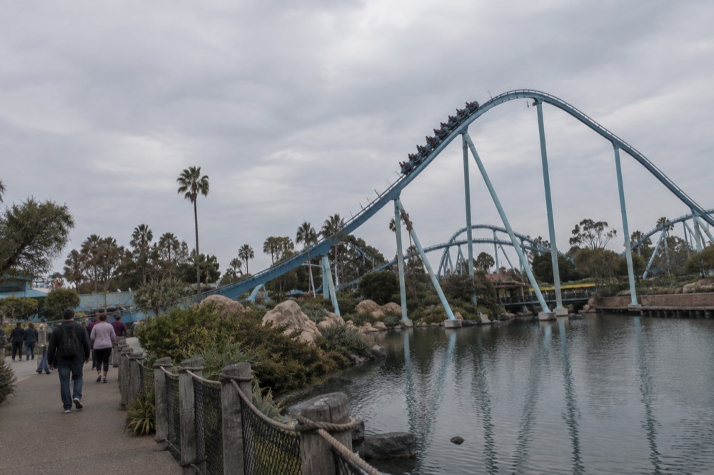 A launch coaster train climbing a track near a park lagoon with visitors below