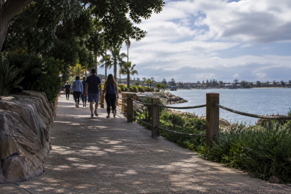 Visitors walking a shaded park pathway between an exhibit building and a lagoon