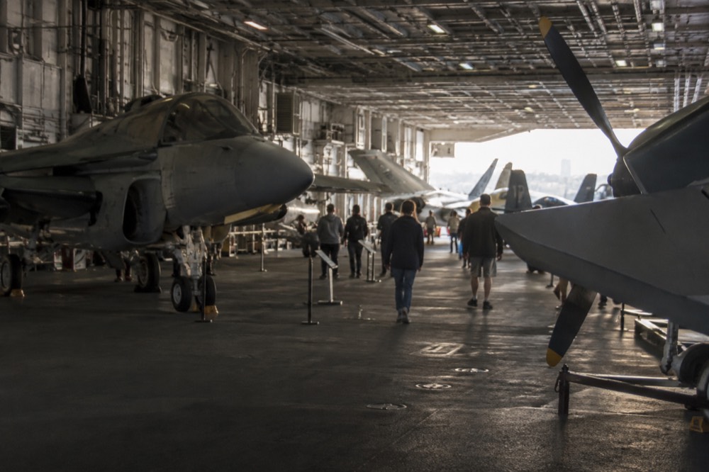 A wide shaded hangar deck interior with aircraft on display and visitors walking between them