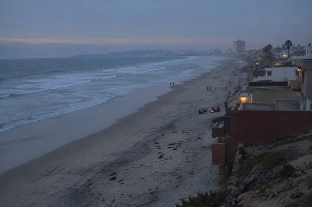 Wide sandy beach with gentle surf and low buildings along the shore under a hazy sky