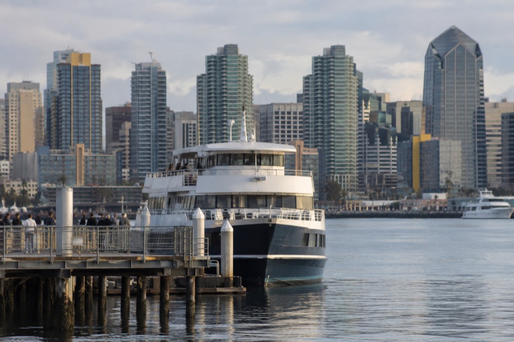 A sightseeing boat at a dock on a calm bay with a city skyline behind it