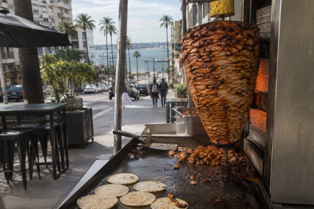 A vertical trompo spit with marinated pork and a grill in front of it