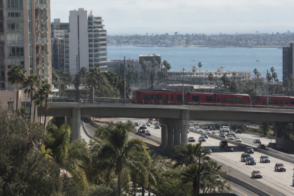 A trolley car crossing a bridge above a San Diego freeway with the bay in the distance