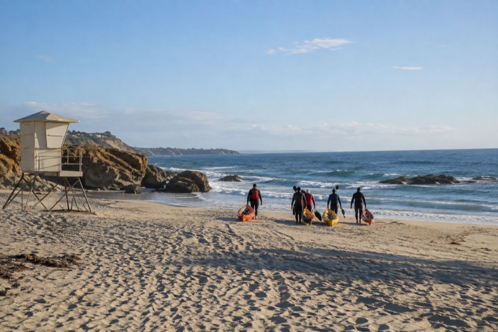 A wide sandy beach with lifeguard tower and a line of paddlers walking kayaks toward the water