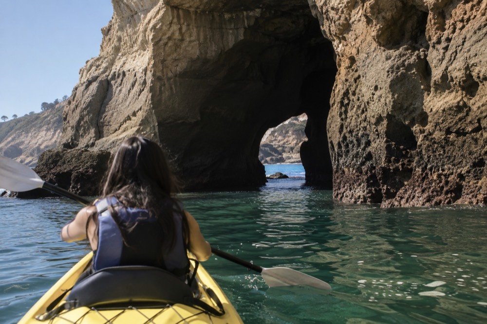 A kayak gliding over clear green water toward a sandstone sea cave opening