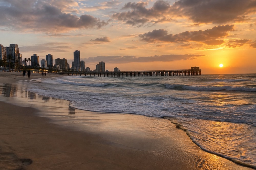 A wide sandy beach with a wooden pier and low breaking waves at golden hour
