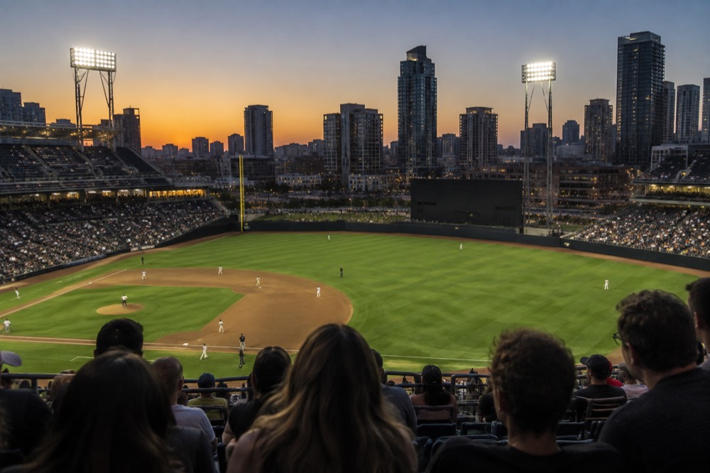 Baseball field at dusk with stadium lights on and a clear sky over the outfield wall