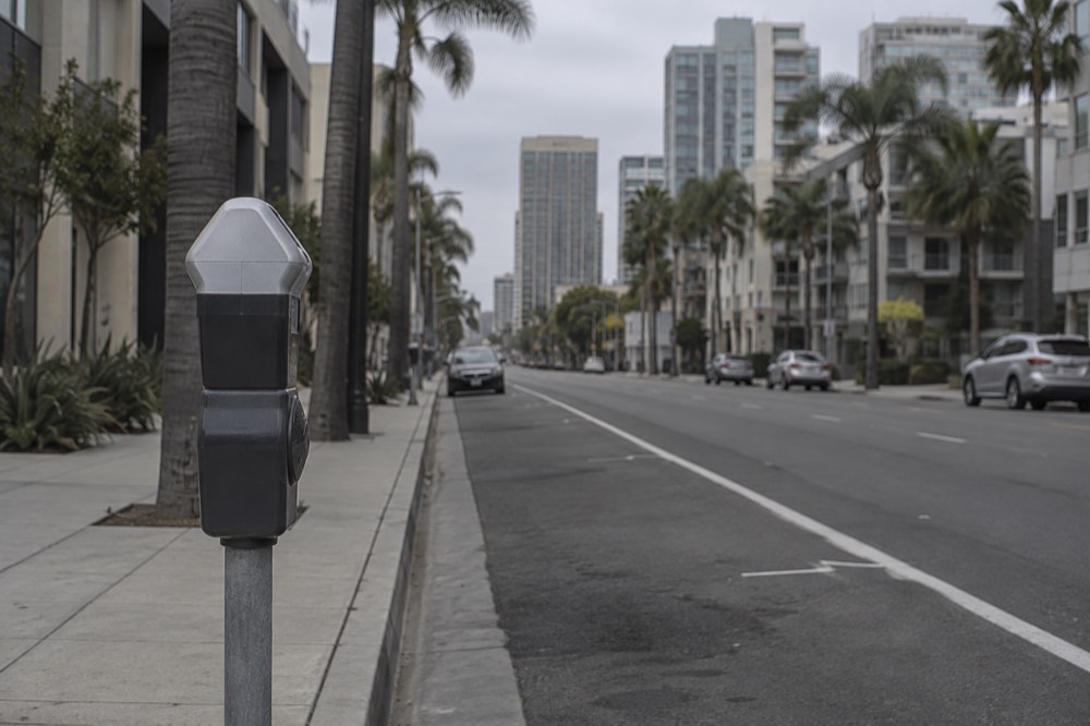 A parking meter next to an empty curbside space on a downtown street with palm trees