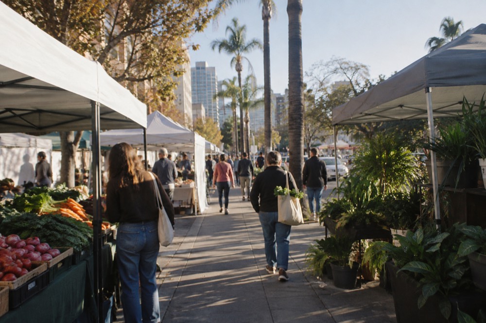 Market stalls lining a tree-lined street with shoppers carrying canvas bags