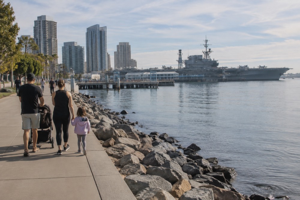 A family walking along the Embarcadero waterfront path with the USS Midway in the distance