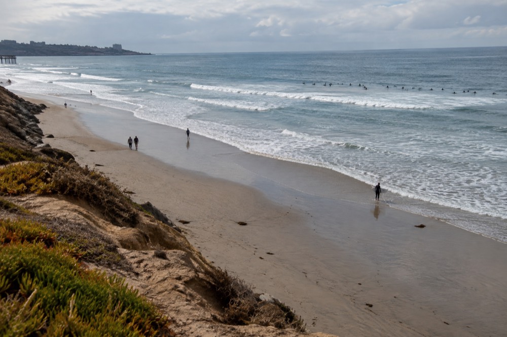 Long sandy beach with gentle rolling waves and surfers sitting on boards beyond the break