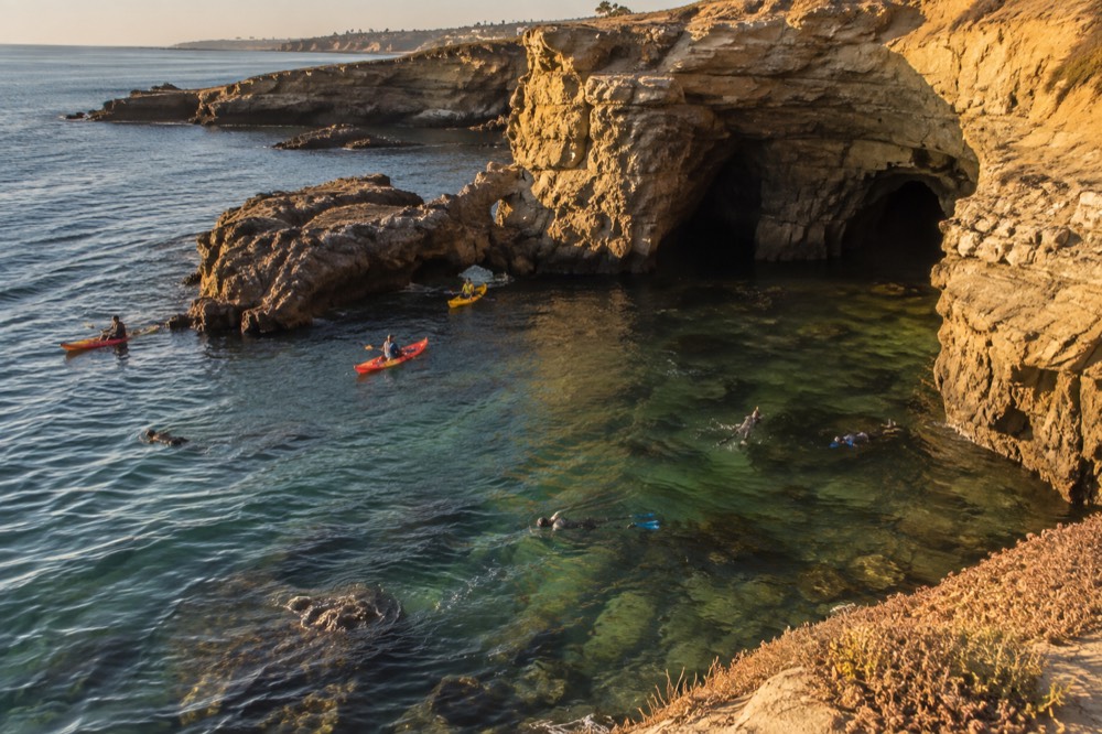 Kayaks and snorkelers in clear green water above a rocky reef near sea caves
