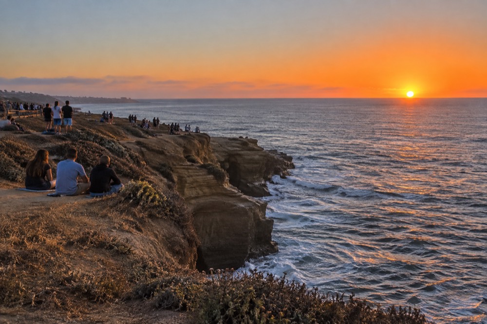 Crowd gathered at Sunset Cliffs watching an orange July sunset over the Pacific