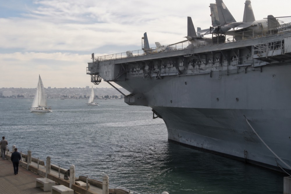 A historic aircraft carrier moored at a downtown waterfront with sailboats passing behind it