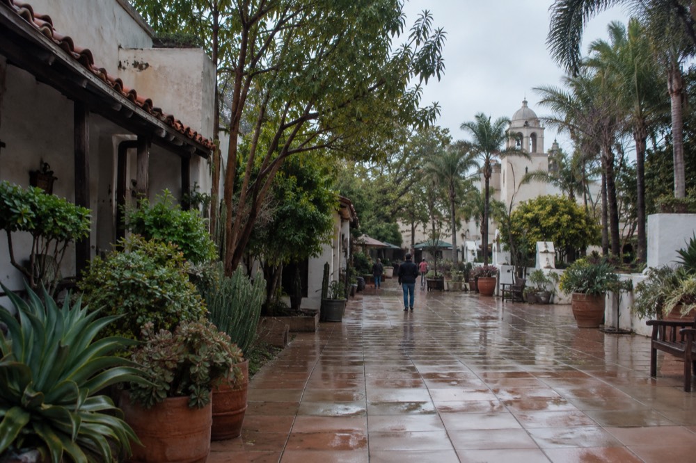 A historic adobe-style plaza with old buildings and leafy trees on an overcast morning
